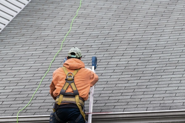 A roofing professional wearing a safety harness climbs a ladder to inspect an asphalt shingle roof before storm season